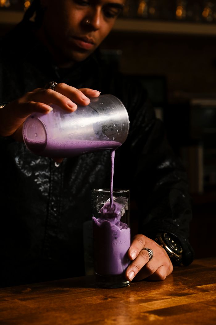 Close-up of a bartender pouring a vibrant purple cocktail in a dimly lit bar.
