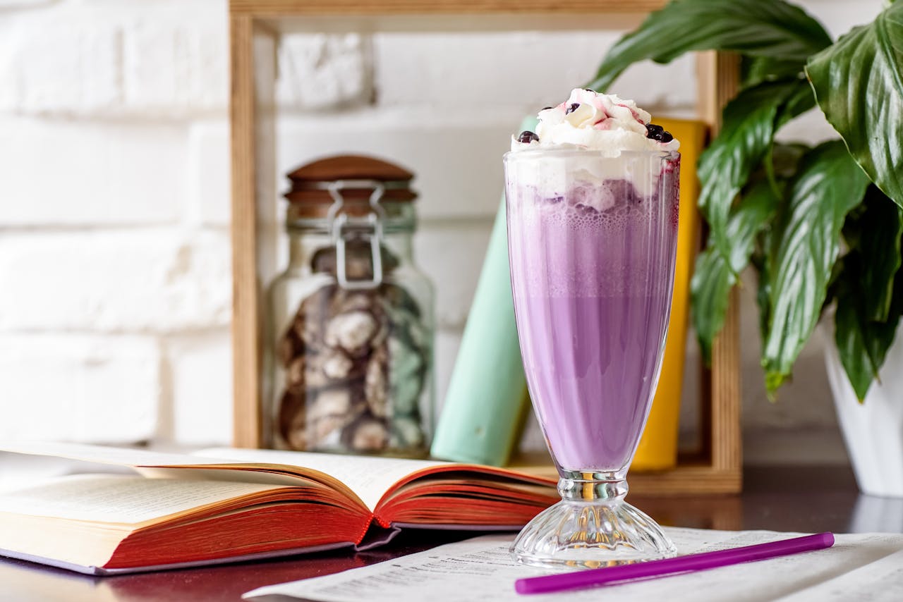 A delicious purple milkshake topped with whipped cream next to open books and a plant.